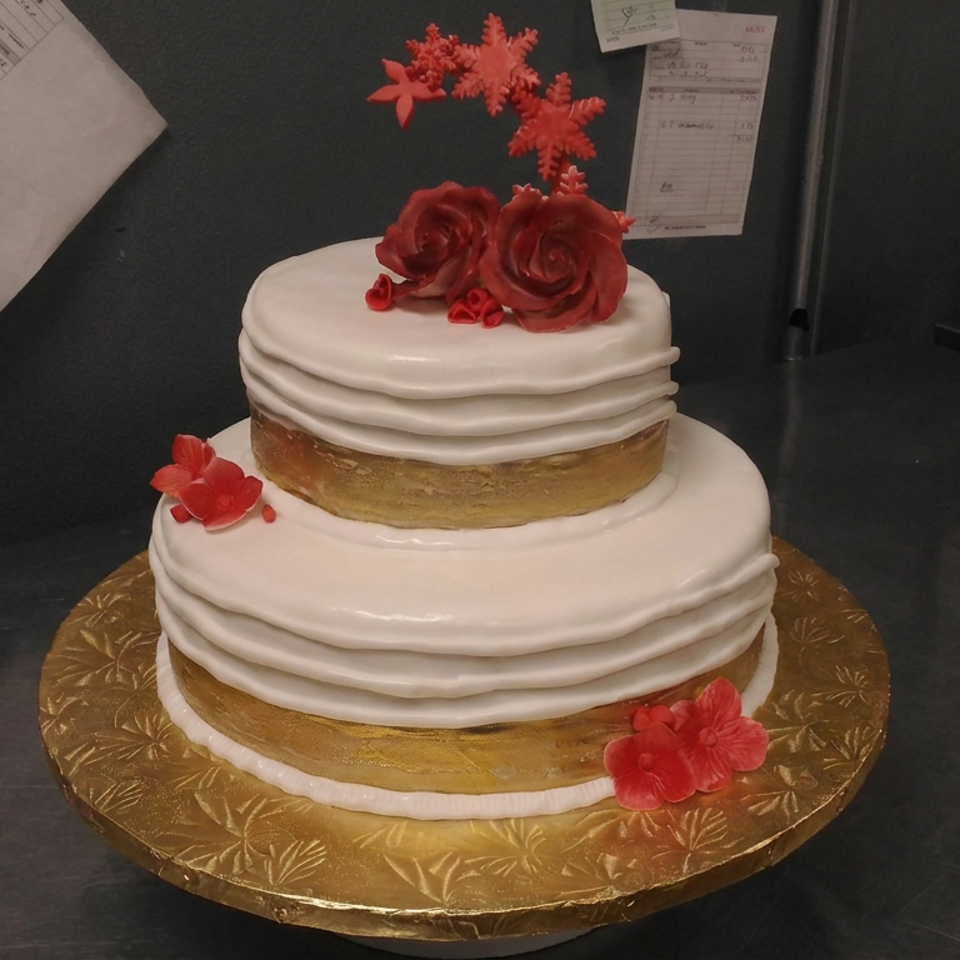 Two-tier white-frosted celebration cake decorated with red roses and flowers on an ornate gold cake board.