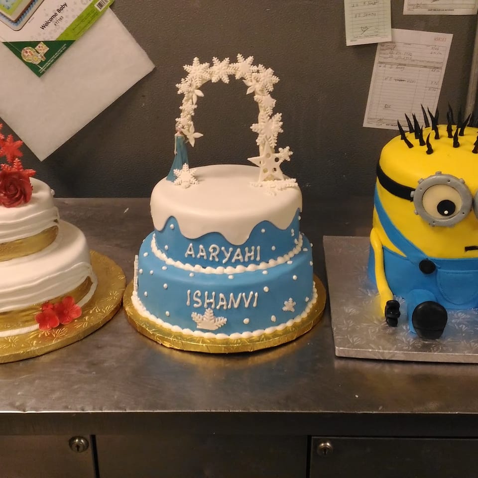 Three decorated cakes displayed on a bakery counter, including floral, snowflake-themed, and Minion designs.